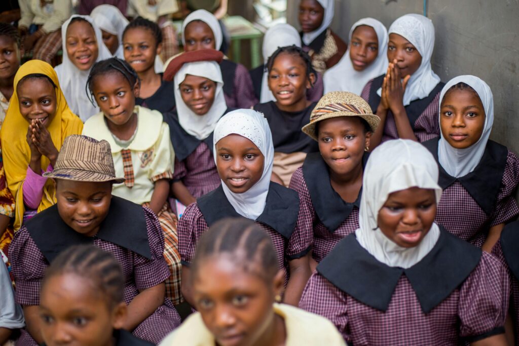 A joyful group of school girls wearing uniforms and headscarves, sitting together and smiling brightly indoors.